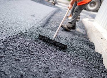 Worker levelling fresh asphalt on a road construction site, industrial buildings and teamwork